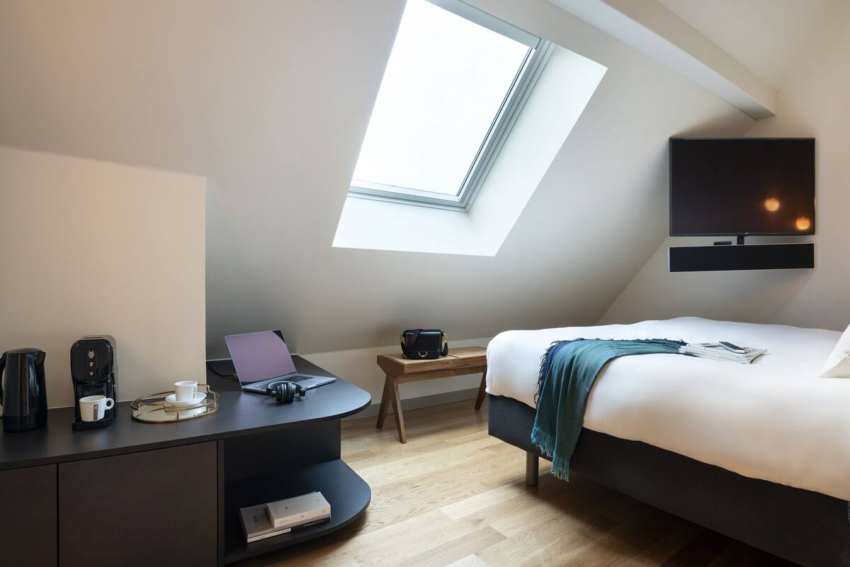 A modern bedroom with a skylight, a bed, a desk with a laptop, a coffee maker, and a TV mounted on the wall.