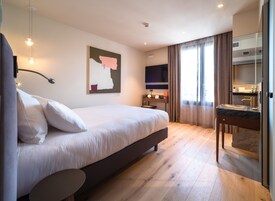 Habitación de hotel moderna con una cama grande, un escritorio con espejo, un televisor de pantalla plana y un ventanal con cortinas.