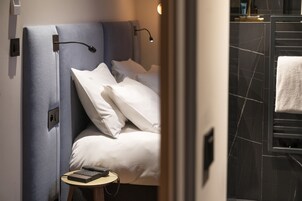 Quarto de hotel moderno com uma cama bem arrumada, uma mesa de cabeceira com um livro e um controle remoto, e um banheiro com porta-toalhas.