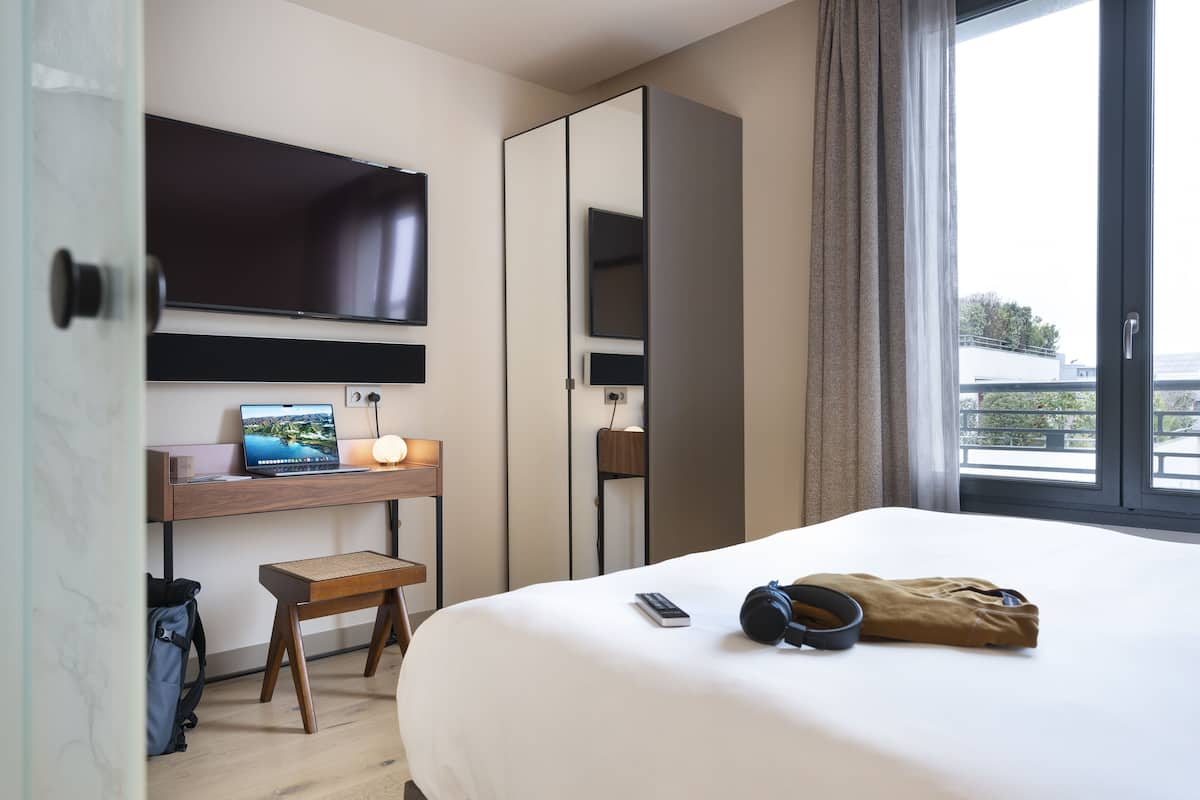 A hotel room with a bed, a desk with a laptop, a chair, a TV mounted on the wall, and a window with a view.
