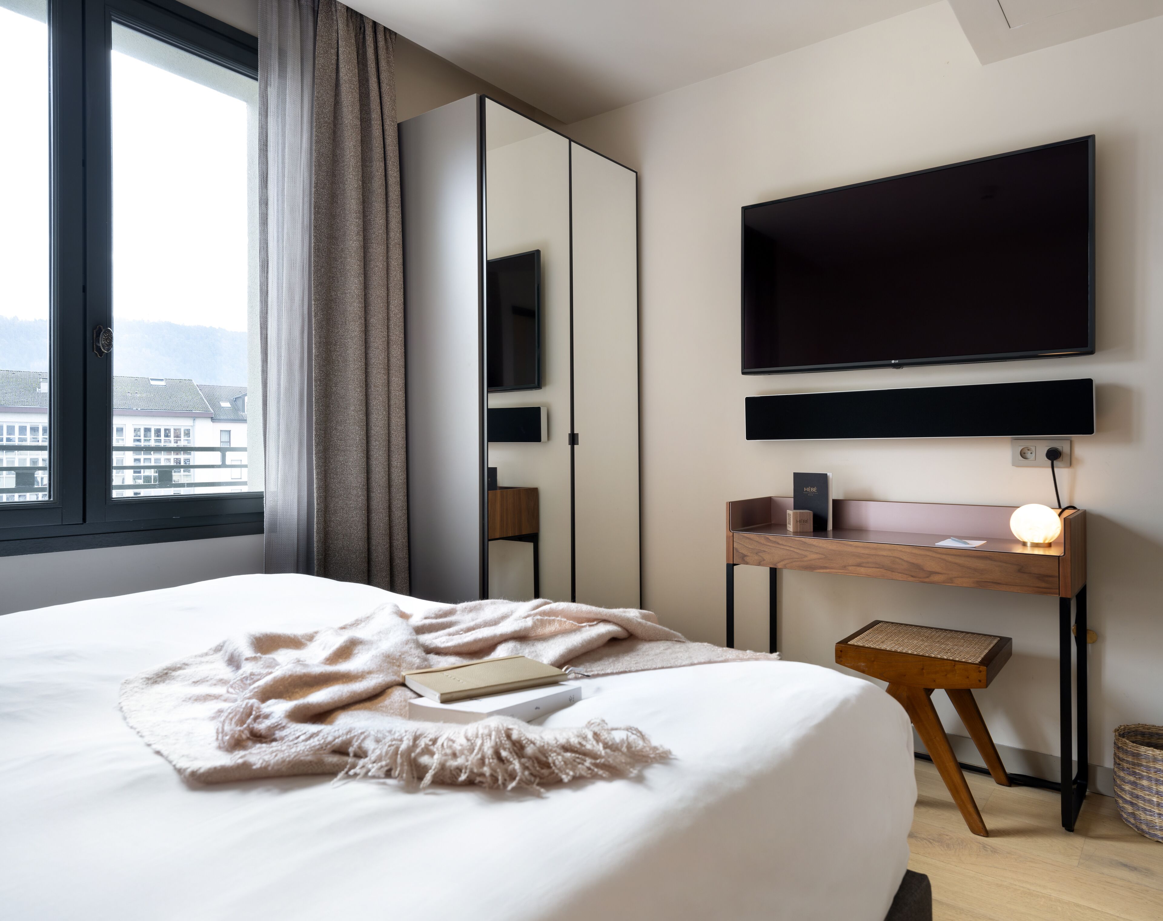 Quarto de hotel moderno com uma cama grande, uma escrivaninha com abajur, televisão de tela plana e vista para edifícios pela janela.
