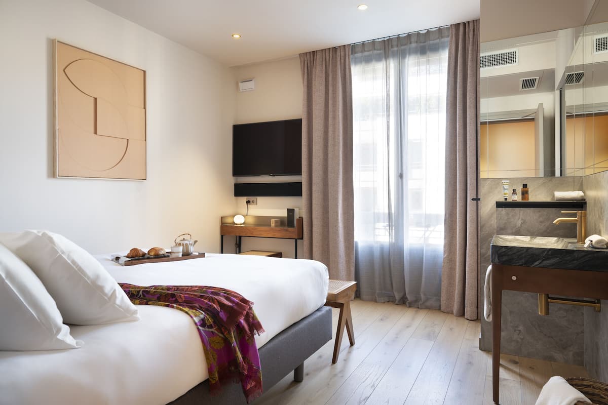 A hotel room with a bed, a desk, a TV, a bathroom with a mirror, and a window with curtains.