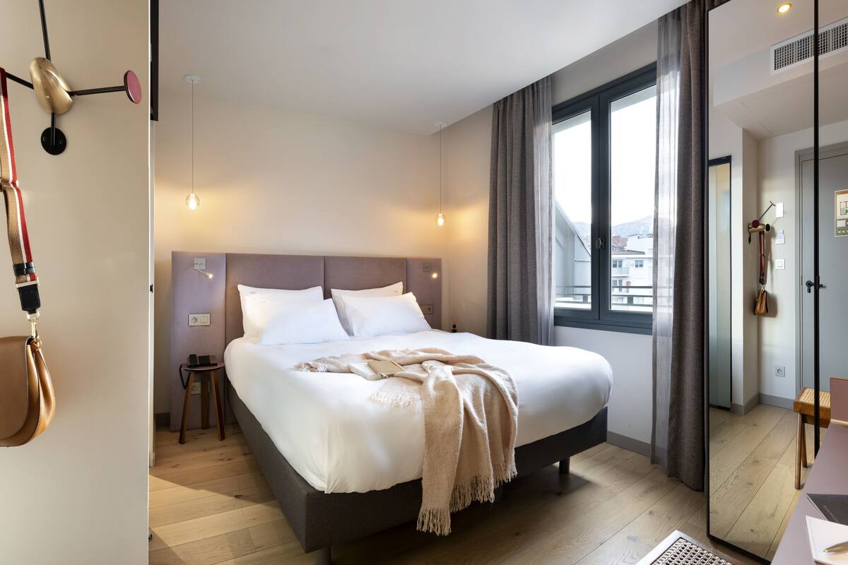 A modern hotel room with a large bed, a bedside table, a lamp, and a view of the city through the window.
