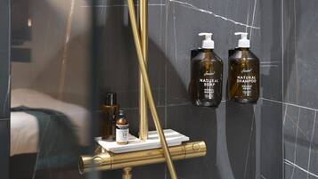 A modern bathroom with a gold showerhead, marble walls, and two bottles of natural shampoo.