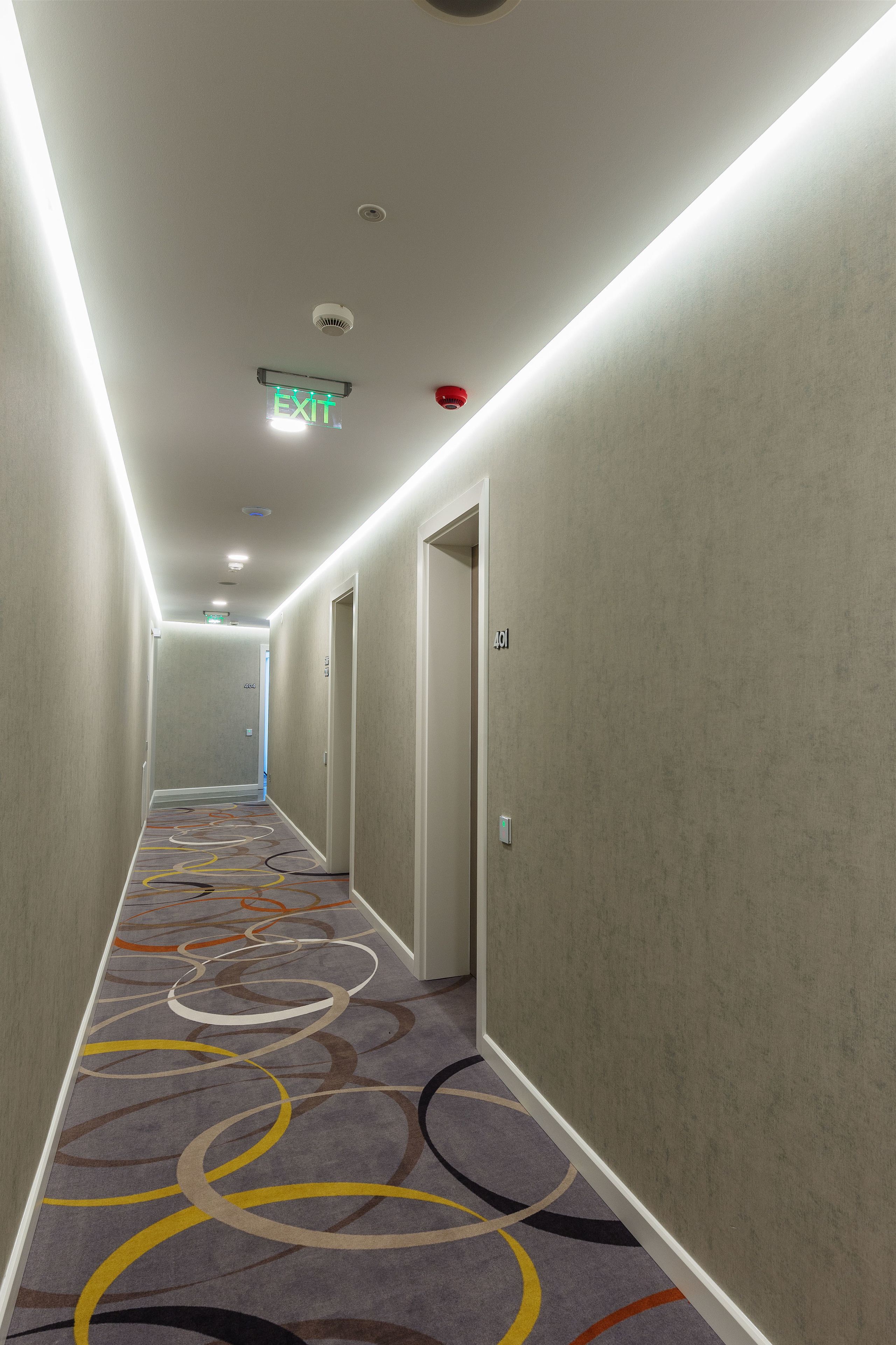 Un pasillo de hotel con alfombra que presenta patrones circulares abstractos, techo con iluminación empotrada y señal de salida.