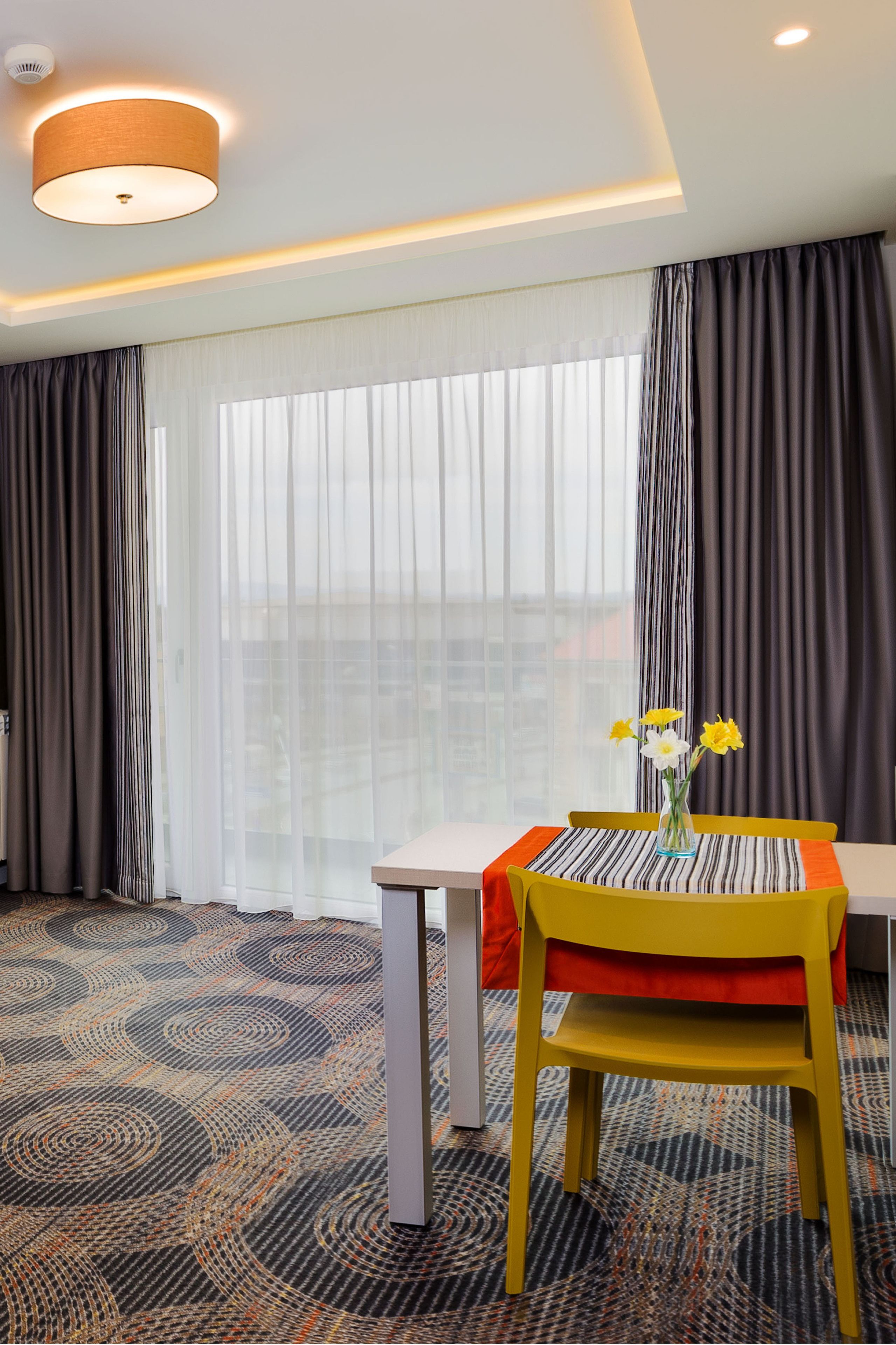 A hotel room with a dining table, a chair, a vase with flowers, and a patterned carpet.