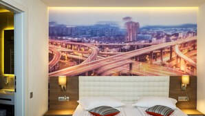 Una habitación de hotel moderna con un mural de paisaje urbano en la pared, un cabecero capitonado y dos lámparas de noche.