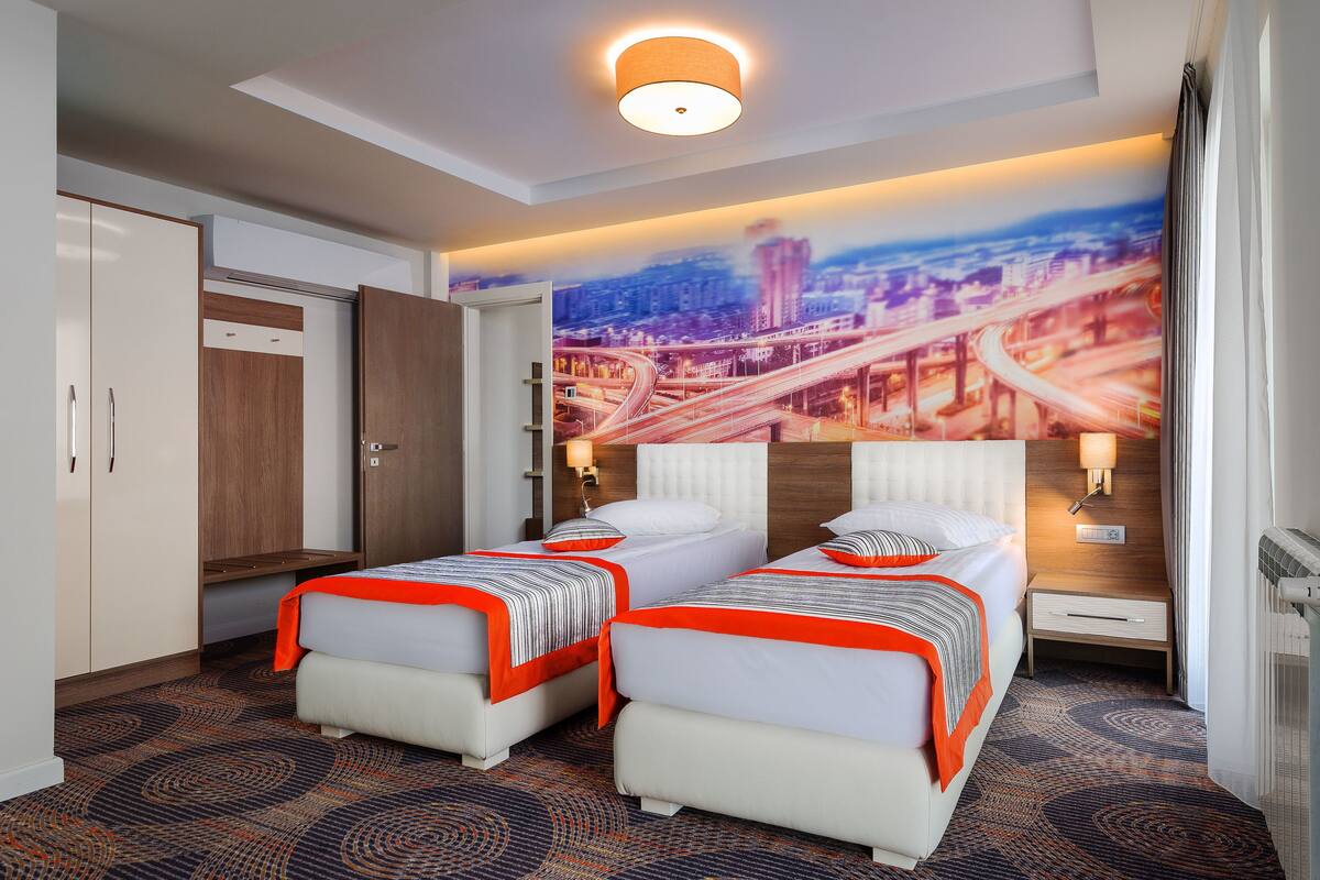 A hotel room with two beds, a cityscape mural, a ceiling light, and a wardrobe.
