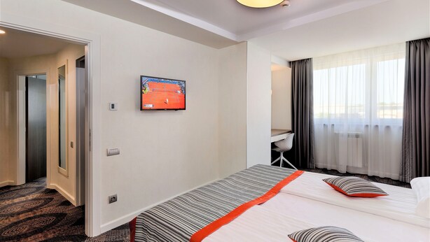 Una habitación de hotel con cama, televisión, escritorio y ventana con cortinas.