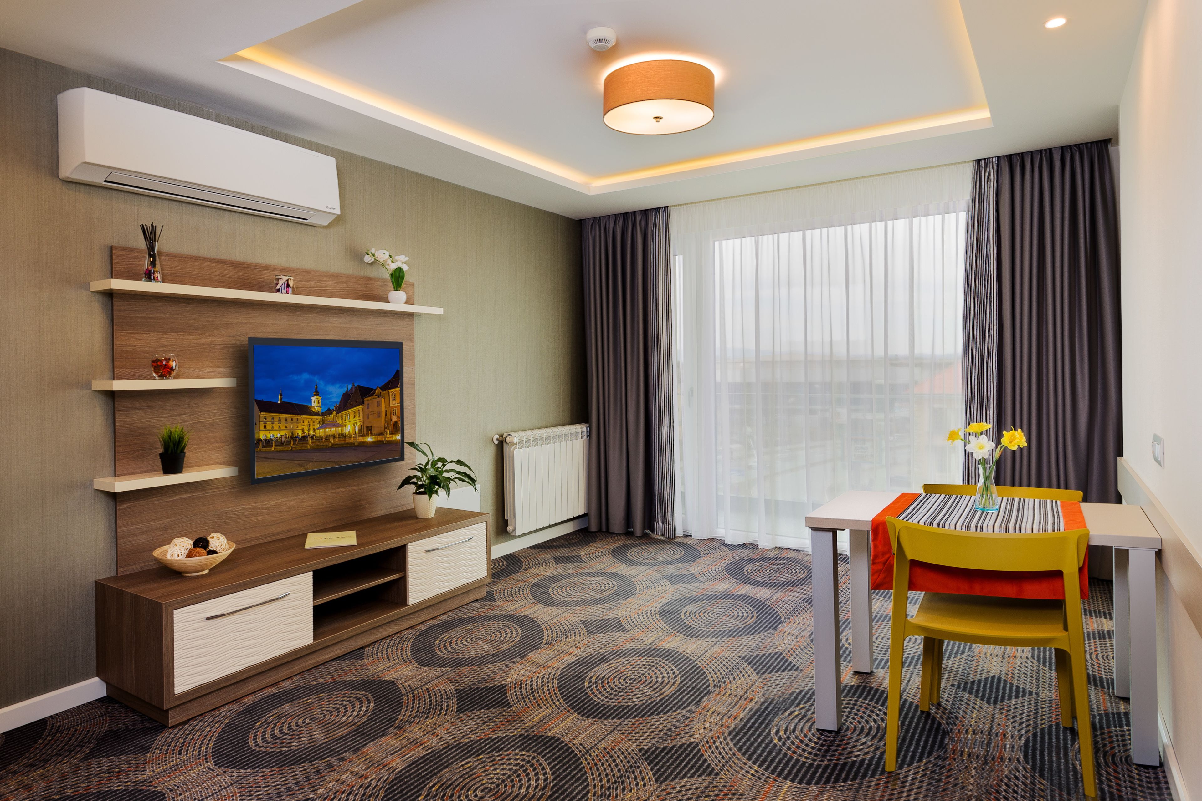 A modern hotel room with a flat-screen TV, a wooden entertainment unit, a dining table with a yellow chair, and a large window with curtains.
