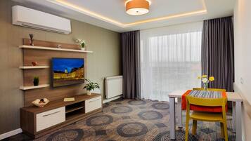 Una habitación de hotel moderna con una televisión de pantalla plana, un mueble de madera para entretenimiento, una mesa de comedor con una silla amarilla y un ventanal con cortinas.