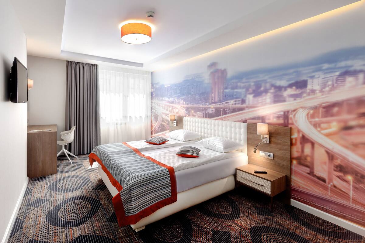 A modern hotel room with a large bed, a desk, and a cityscape mural on the wall.