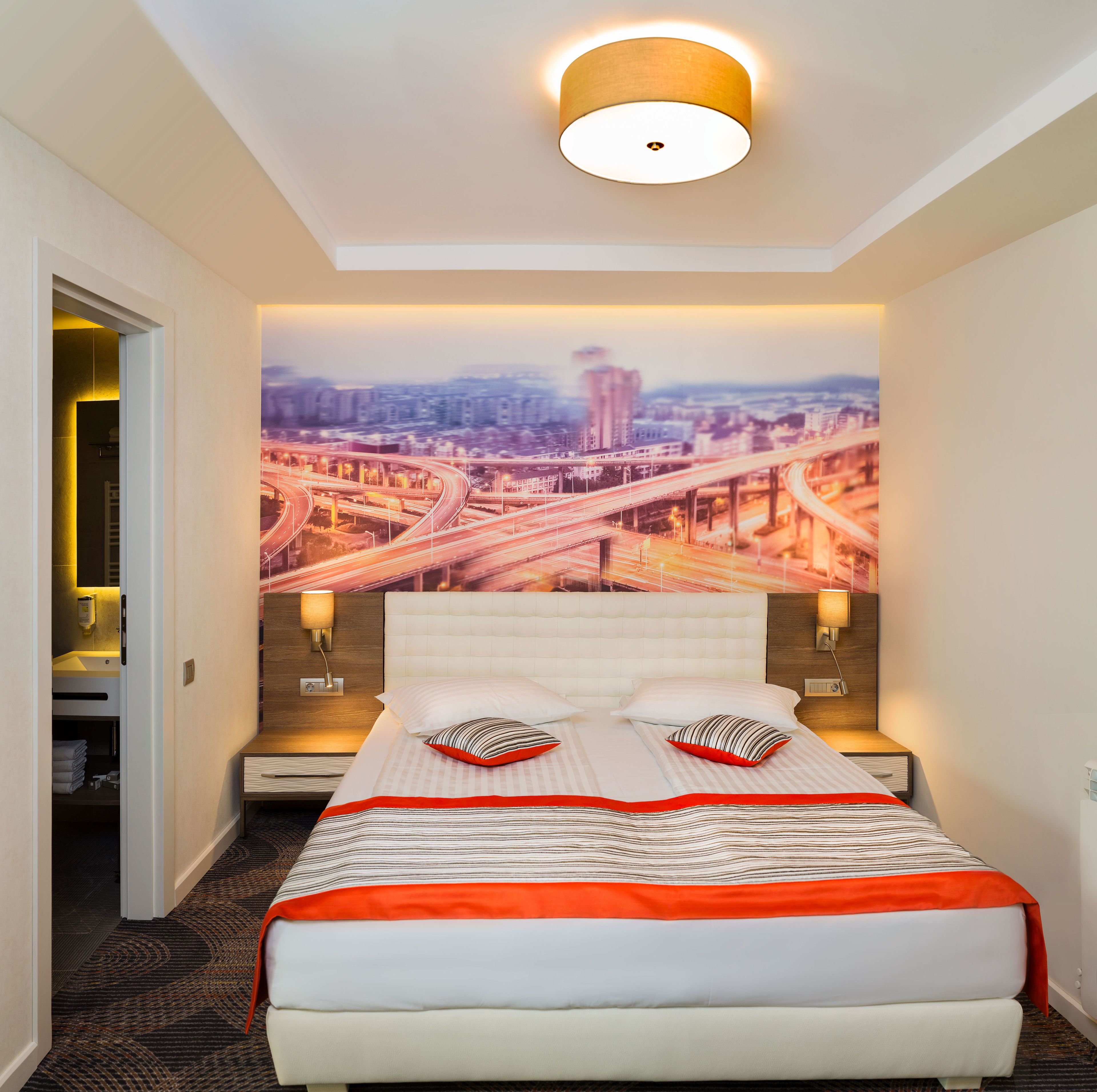 A hotel room with a large bed, two bedside tables, a headboard, a wall-mounted light fixture, and a cityscape mural on the wall.