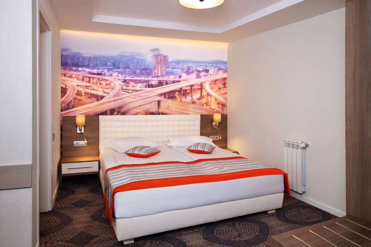 A hotel room with a large bed, a cityscape mural on the wall, a nightstand, and a radiator.