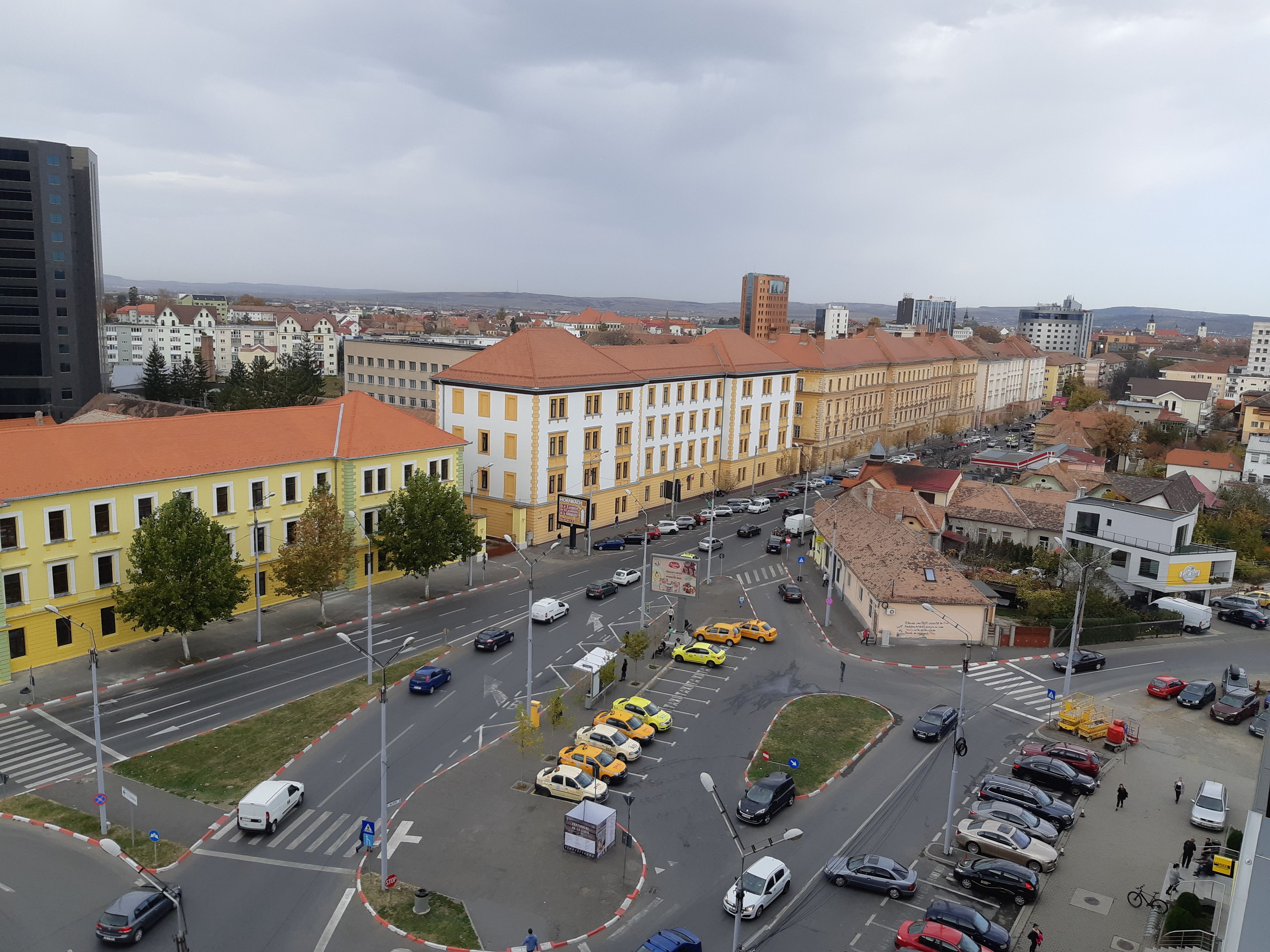 A cityscape with a prominent yellow building with a red roof, surrounded by other buildings and a busy street with cars and pedestrians.