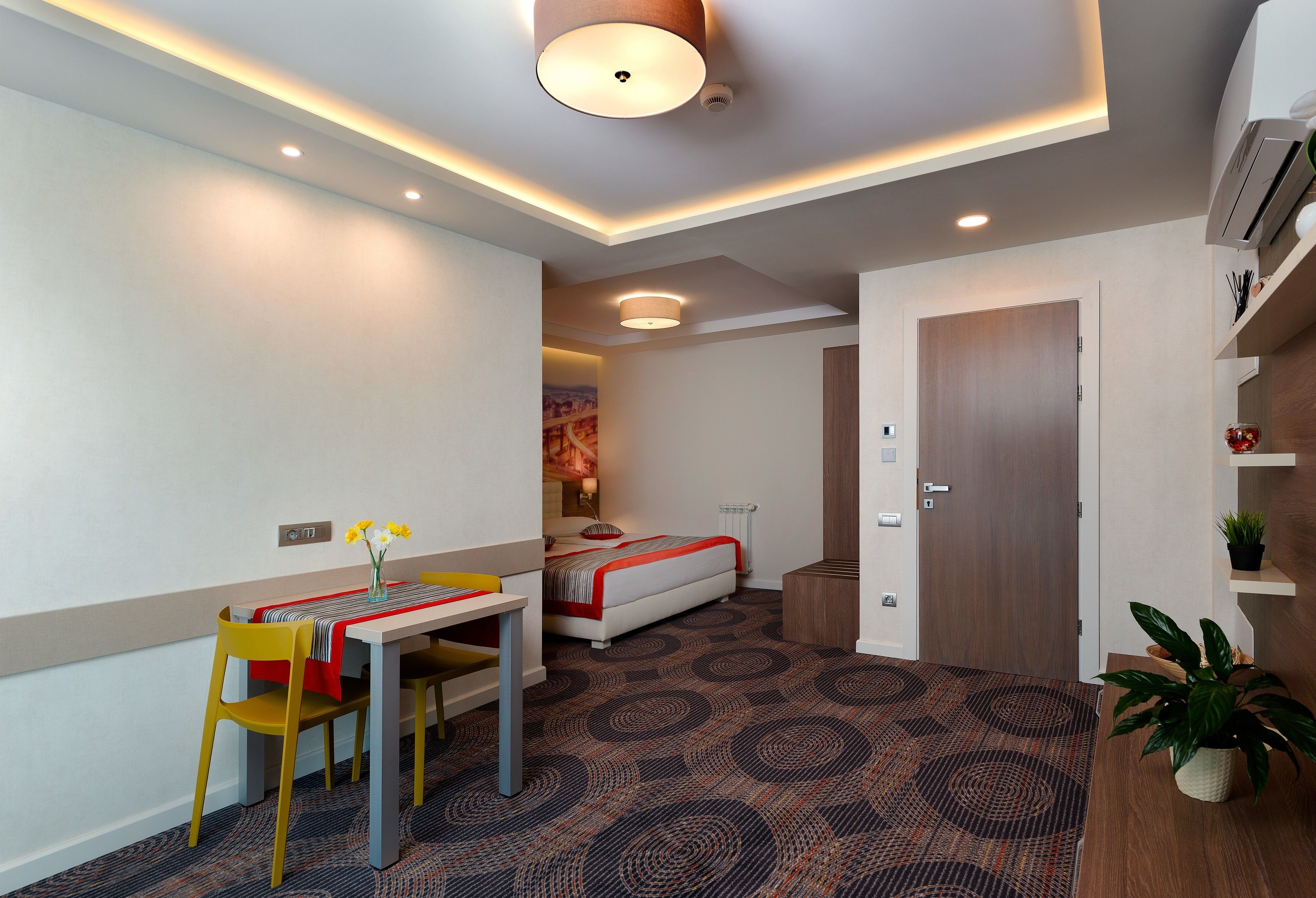 A modern hotel room with a bed, a desk with a chair, a small table with a vase of flowers, and a potted plant.