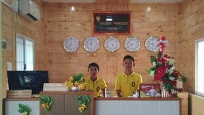 Un comptoir d’accueil à l’hôtel Golden Princess, avec des horloges indiquant différents fuseaux horaires, une enseigne indiquant « Réception » et des employés portant des uniformes jaunes.