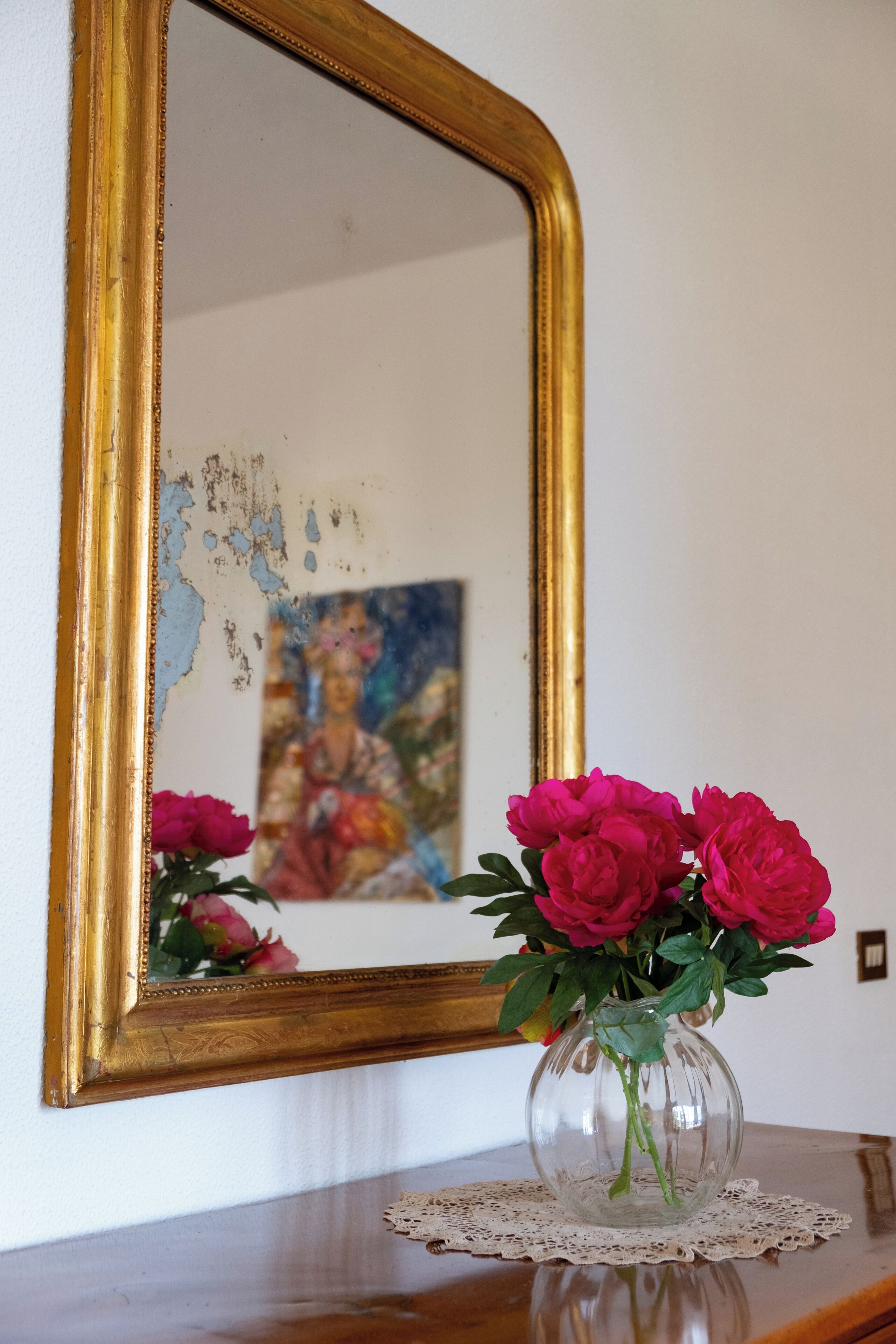 A vase with flowers on a wooden table, a gold-framed mirror reflecting a painting, and a lace doily underneath the vase.