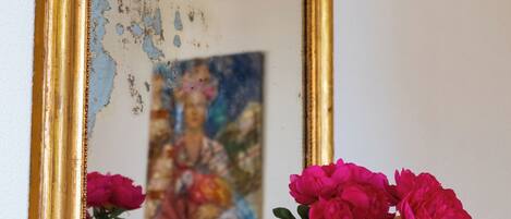 A vase with flowers on a wooden table, a gold-framed mirror reflecting a painting, and a lace doily underneath the vase.