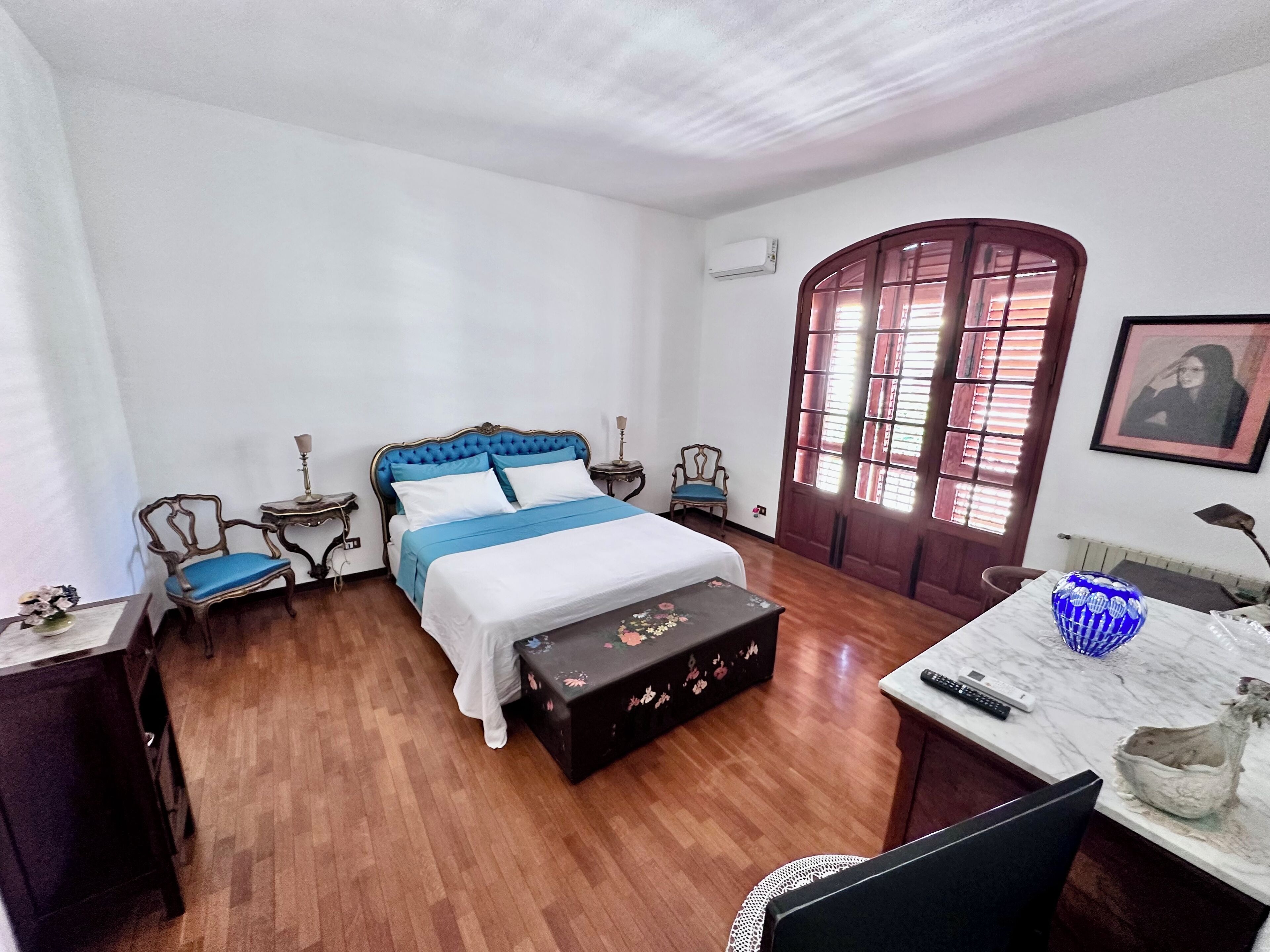 Un dormitorio con cama, suelo de madera, un escritorio con silla, una mesita pequeña, una ventana con contraventanas y un cuadro enmarcado en la pared.