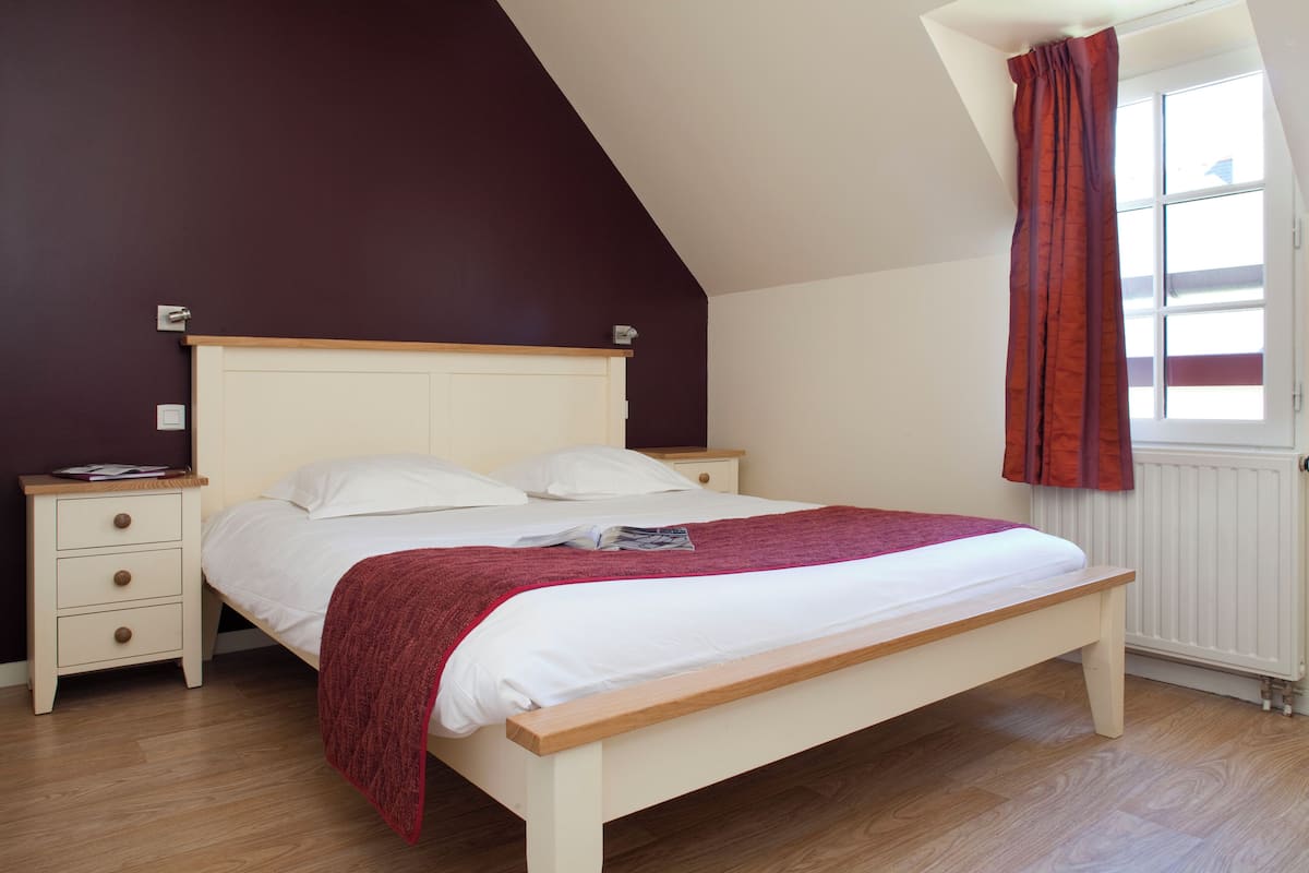 Une chambre à coucher avec un lit, des tables de chevet, une fenêtre avec des rideaux et un plafond en pente.