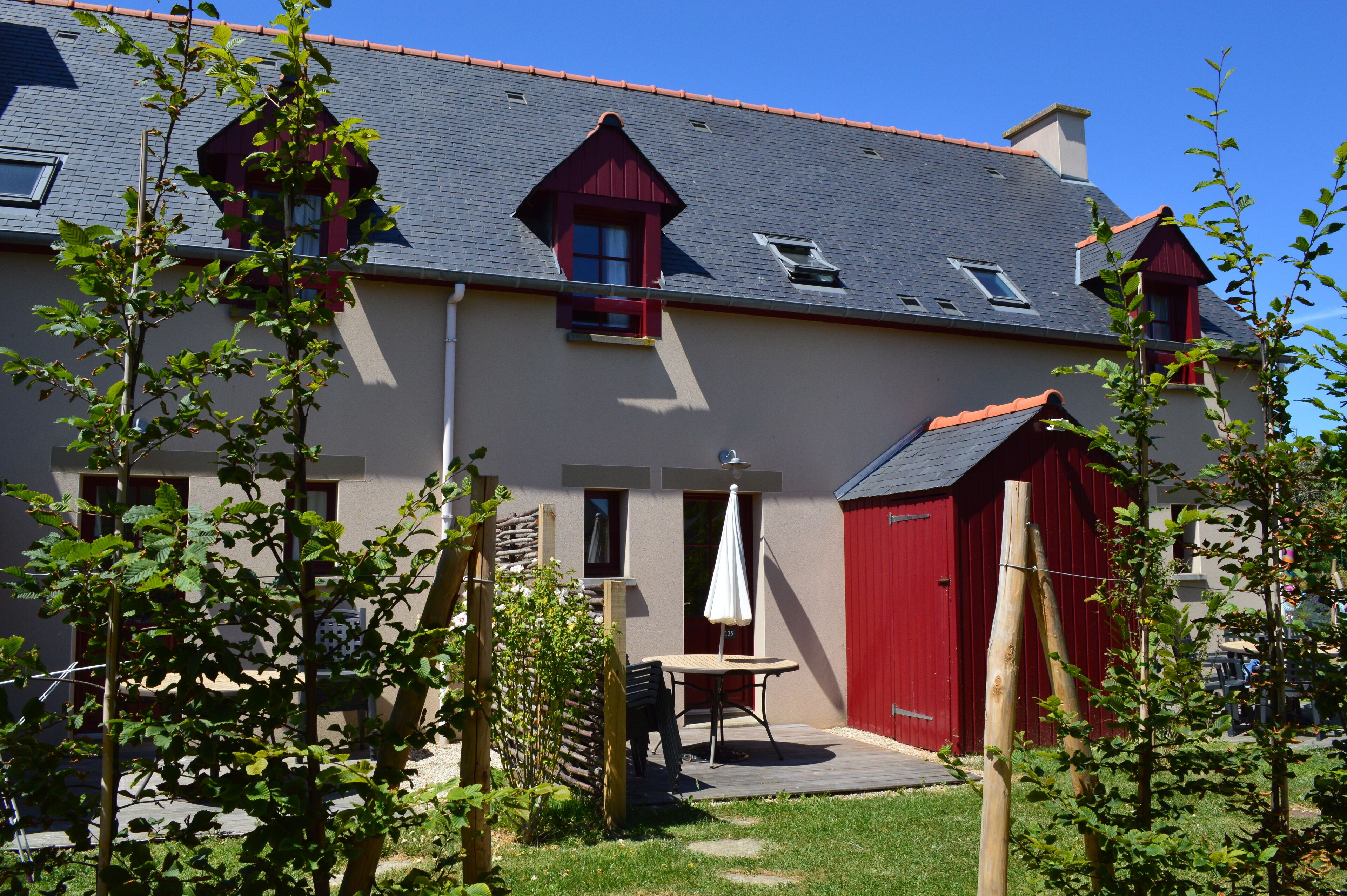 Un bâtiment à deux étages, avec une structure ressemblant à une grange peinte en rouge, un petit patio avec une table et des chaises, et un jardin agrémenté d’arbres et d’arbustes.