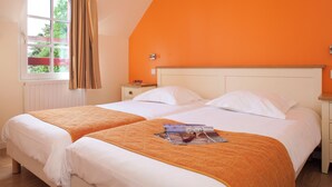 A double bed with white linens and an orange throw blanket, a nightstand with a lamp, and a window with curtains. - Le Village Cancalais (Cancale)