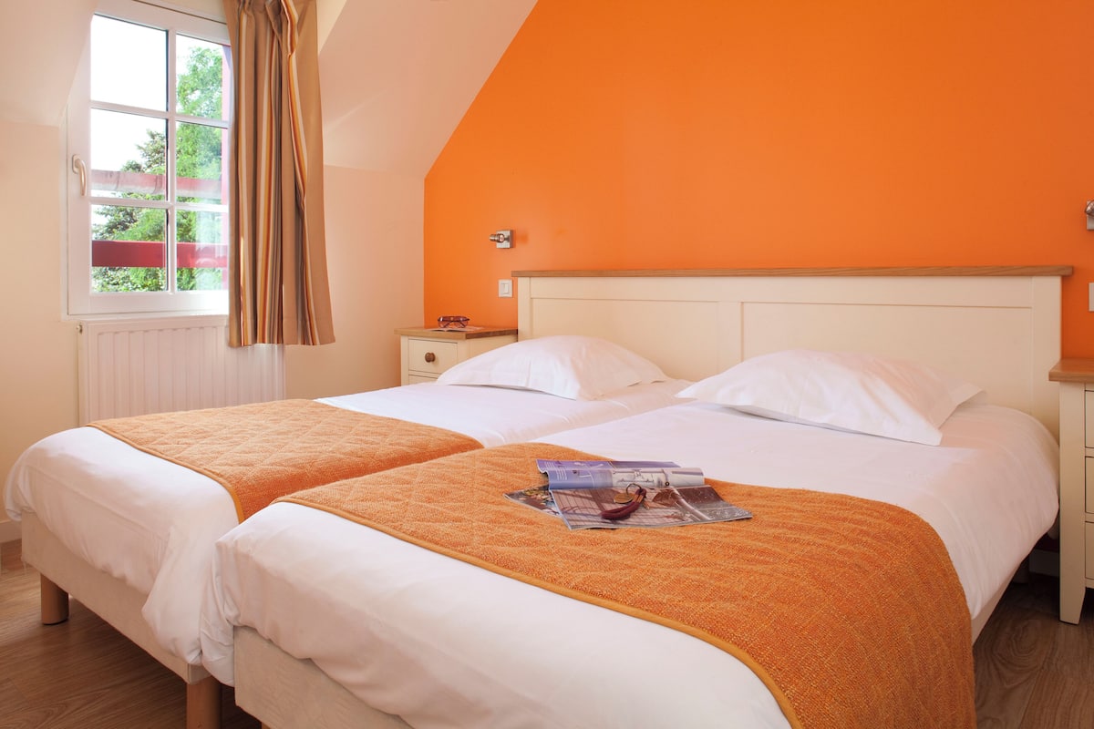 Un lit double avec du linge de lit blanc et un plaid orange, une table de chevet avec une lampe, et une fenêtre avec des rideaux.