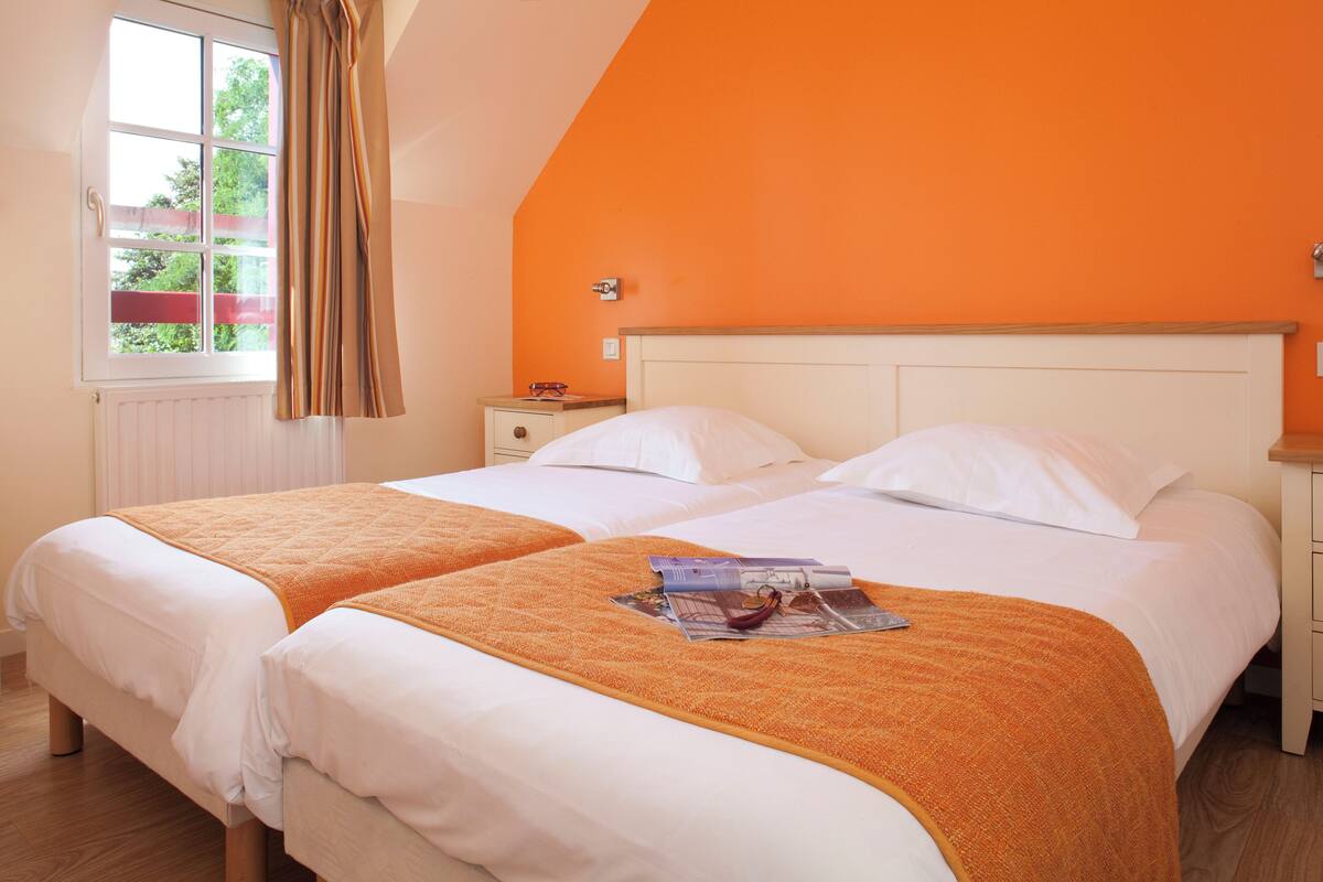 Una cama doble con ropa de cama blanca y una manta naranja, una mesita de noche con lámpara y una ventana con cortinas.