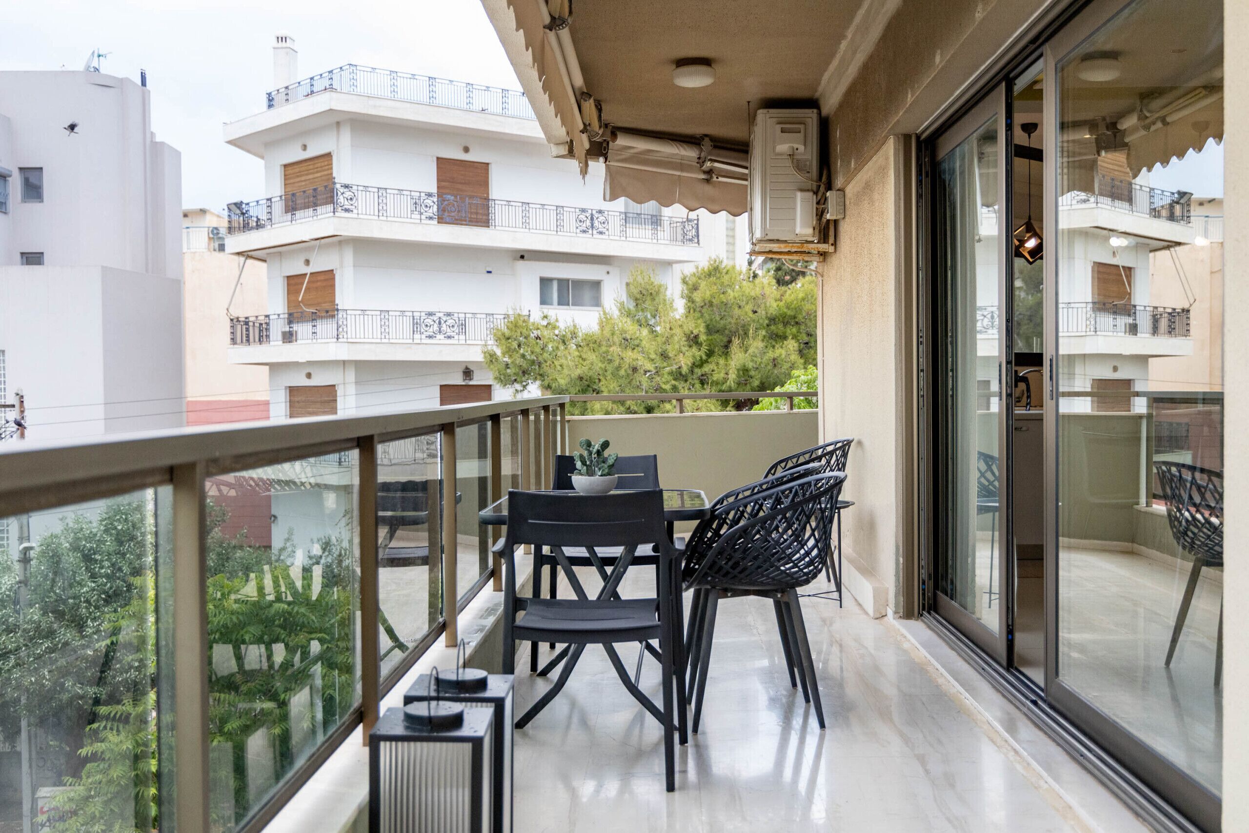 Un balcon avec une table et des chaises, un pot de fleurs et une vue sur les bâtiments et les arbres voisins.