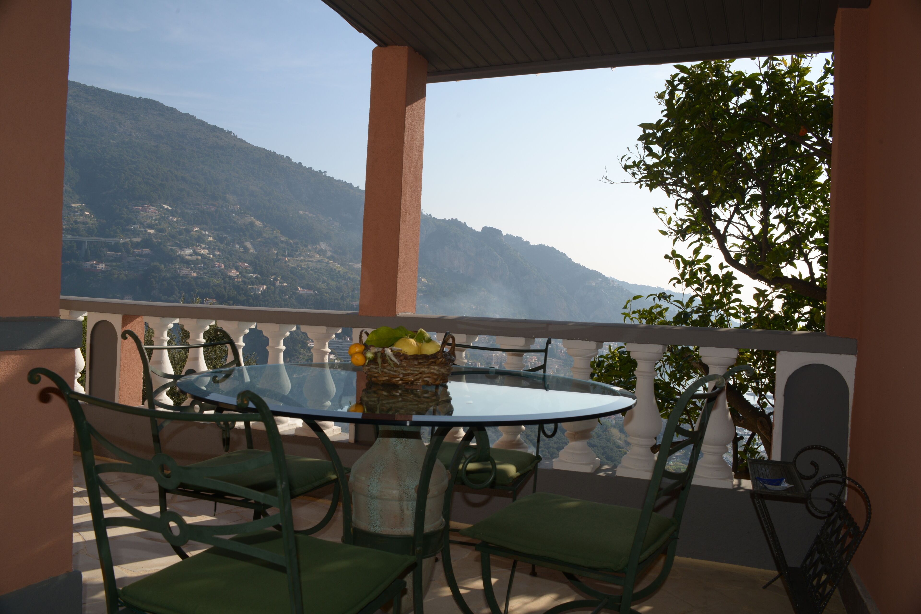 A round glass table with chairs, a wicker basket with fruits, and a view of a mountainous landscape.