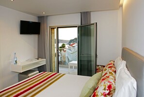 A hotel room with a bed, a flat-screen TV, a desk with a bottle and glasses, and a view of the outside.