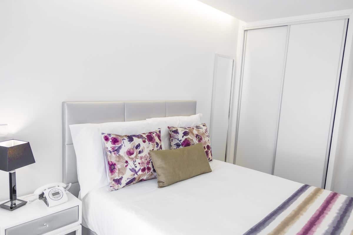 Una cama bien hecha con almohadas florales, una lámpara de noche y un teléfono sobre una mesita blanca.