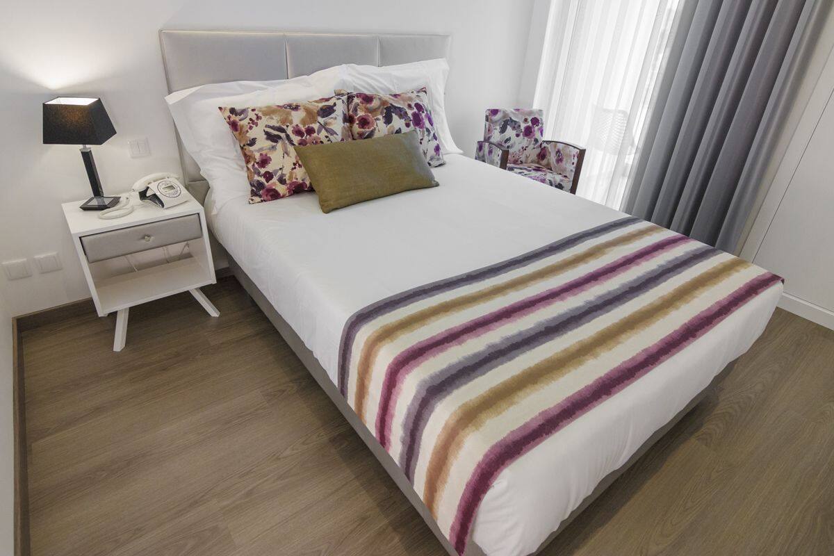 Una cama bien hecha con una colcha a rayas, una mesita de noche con una lámpara y una silla con tapicería floral.