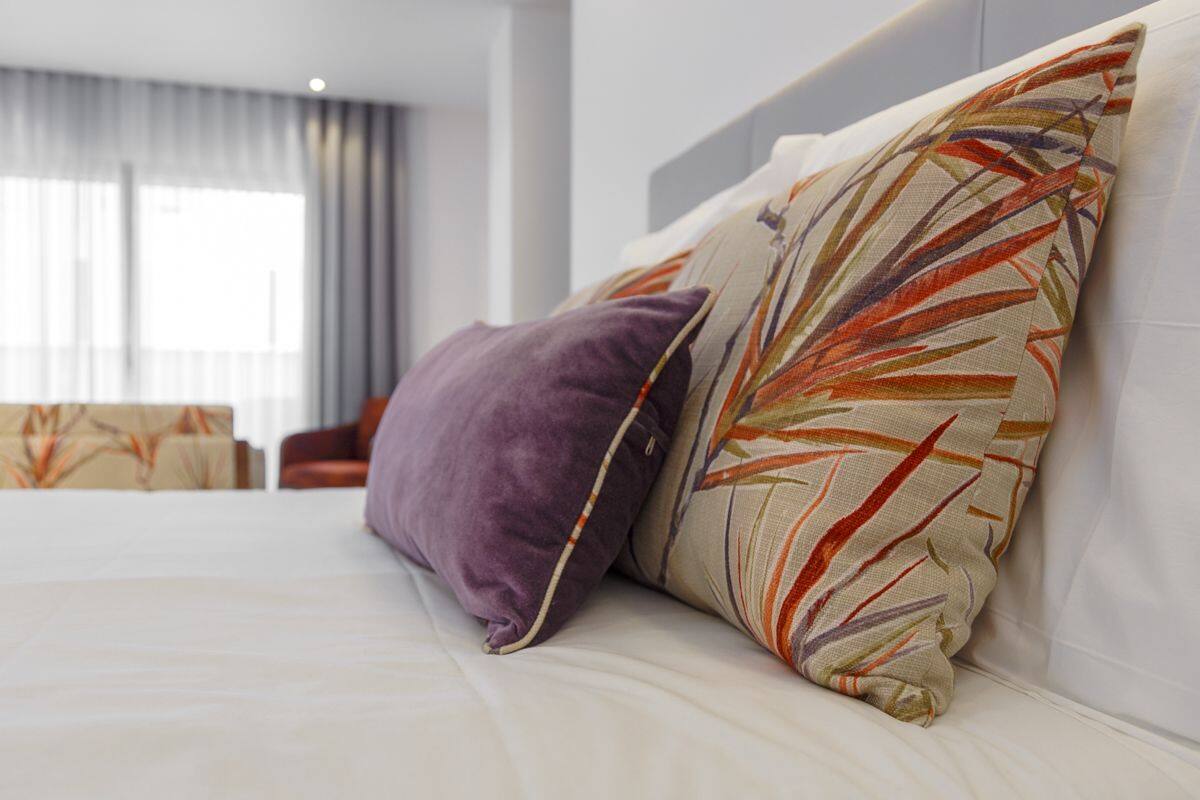 Una cama bien hecha con dos almohadas, una morada y otra con estampado de hojas, en una habitación de hotel moderna con un ventanal grande y cortinas.