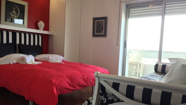Habitación de hotel con cama roja, cabecera blanca y vista al mar a través de un ventanal grande.