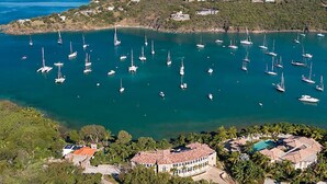 Vista aérea de una zona costera con numerosos veleros amarrados en una bahía. Se observan varios edificios con tejados de tejas rojas, rodeados de vegetación y colinas al fondo.