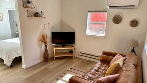 Una sala moderna con un sofá de cuero, un televisor de pantalla plana, un mueble bajo para la televisión de madera, una mesita con un florero y una ventana con persianas.