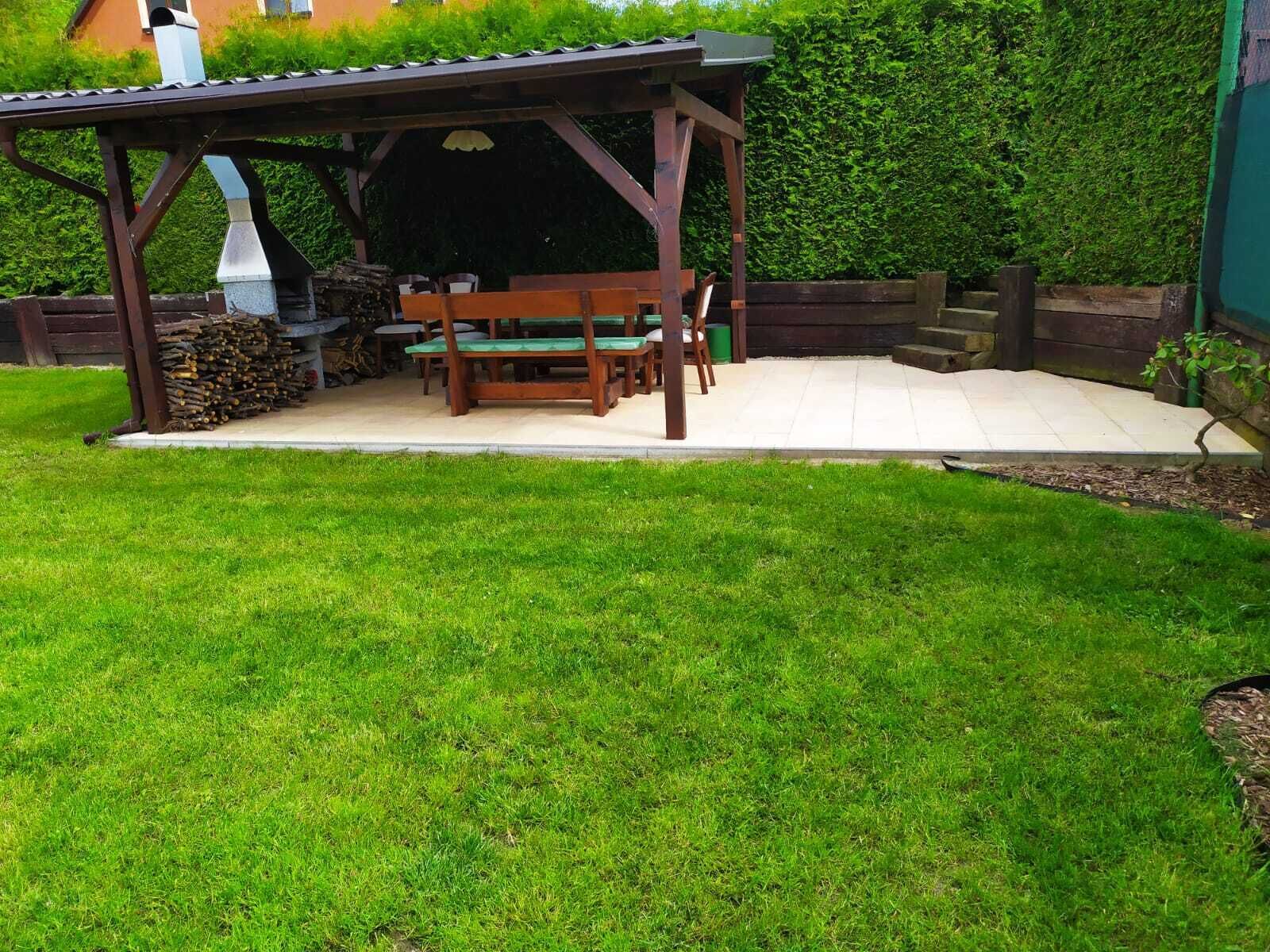 A wooden pergola with a bench and a table, surrounded by a well-maintained lawn and a hedge.