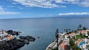 A coastal view with a rocky shoreline, a clear sky, and a resort area with buildings and palm trees. - Spacious, light & cozy apartments with ocean views (Santiago del Teide)