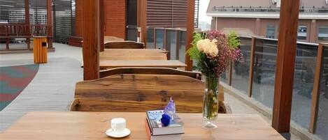 Une table en bois avec une tasse et un livre, un vase rempli de fleurs et un banc dans un espace extérieur abrité, d’où l’on peut admirer les bâtiments.