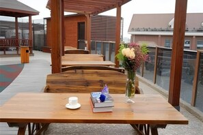 A wooden table with a cup and a book, a vase with flowers, and a bench in a covered outdoor area with a view of buildings.