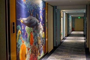 Pasillo de hotel con un mural de una ballena y peces tropicales, piso alfombrado y puertas que dan a las habitaciones.