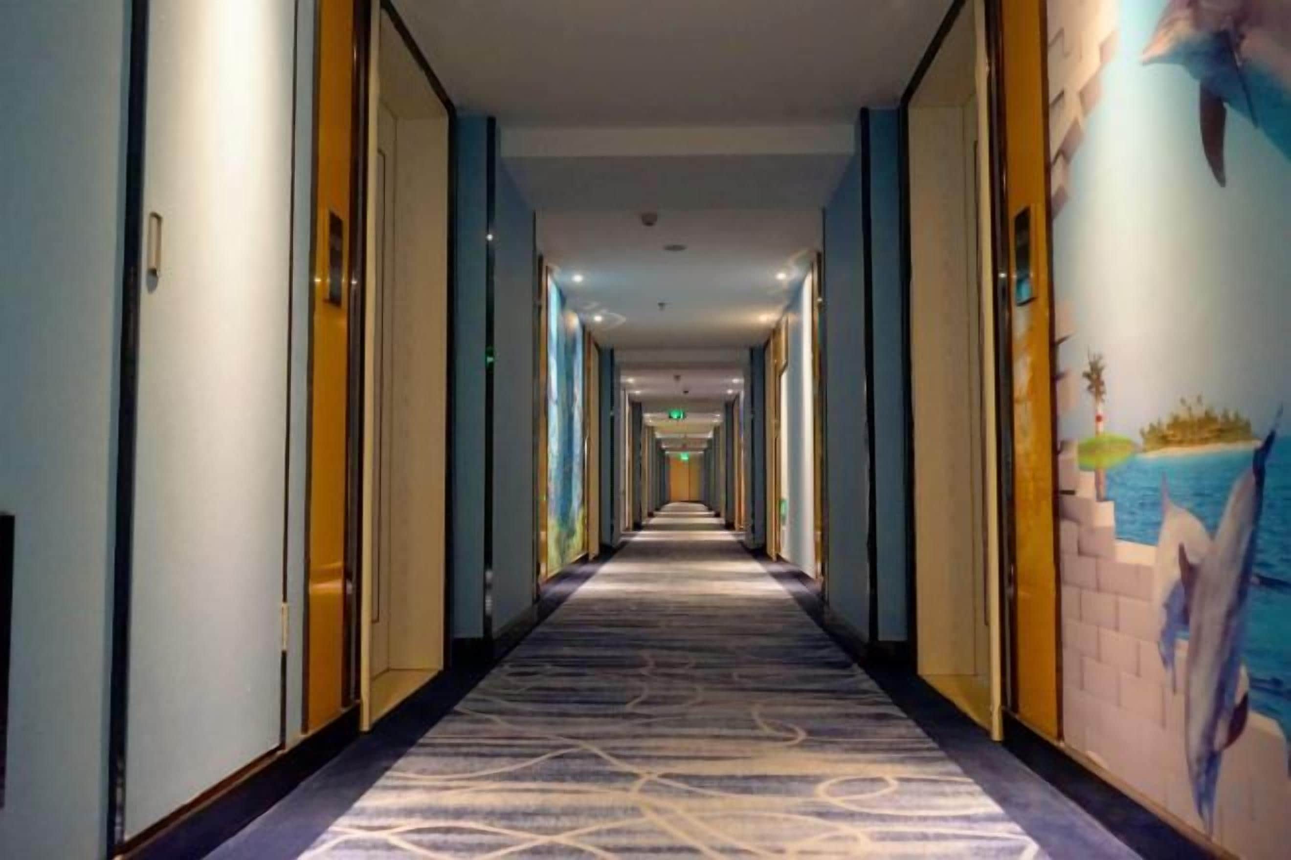 Corredor de hotel com piso revestido de carpete, portas em ambos os lados e um mural na parede da direita.