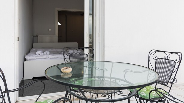Une table à plateau en verre, des chaises en métal, un lit avec du linge de maison blanc et un radiateur dans une pièce aux murs blancs.