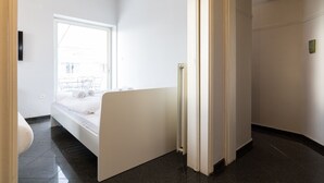 Une chambre d’hôtel moderne avec une grande fenêtre, un lit, un radiateur et une porte donnant sur une autre pièce.