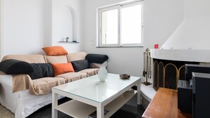 Un salon moderne avec une table basse en verre, un canapé beige agrémenté de coussins orange et noirs, une cheminée blanche et une fenêtre avec des stores.