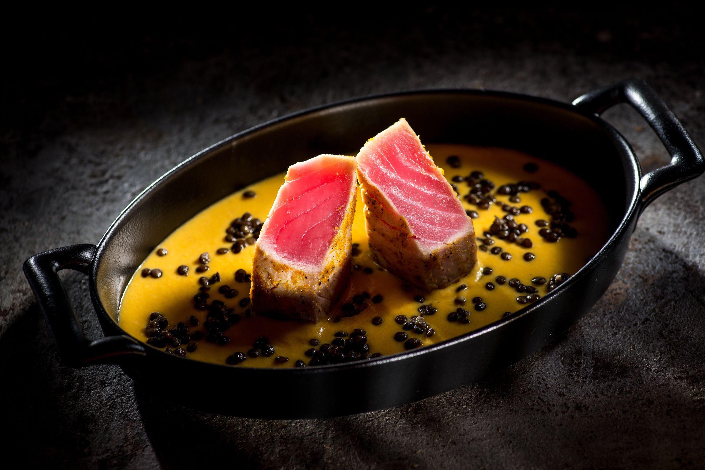 A skillet with two pieces of meat and black peppercorns.