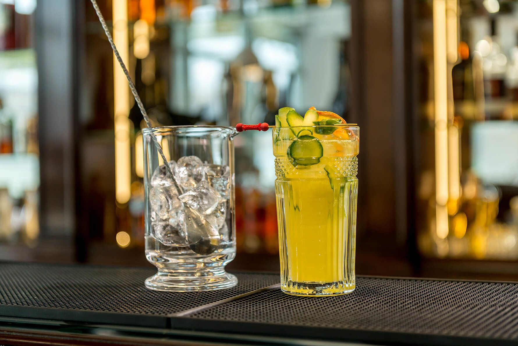 Two glasses of beverages on a bar counter, one with ice and the other with fruit garnishes.