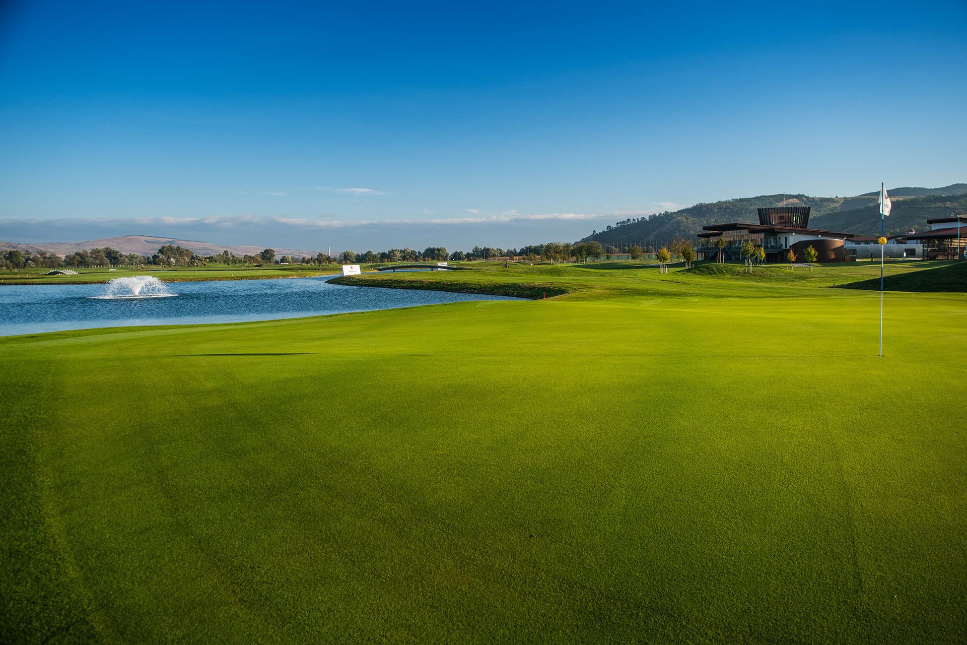 A golf course with a water hazard, a flagstick, and a building with a modern design in the background.